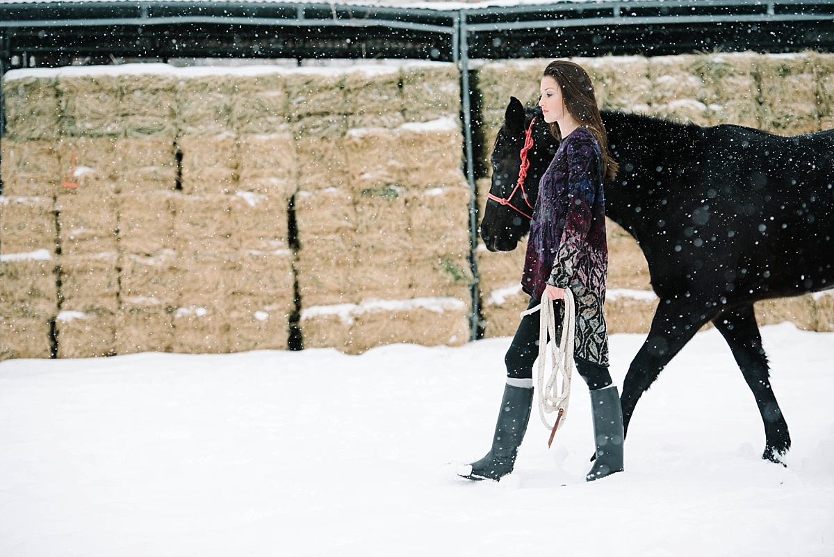 girl walking her black horse in the snow in front of a stack of hay, utah equestrian photographer
