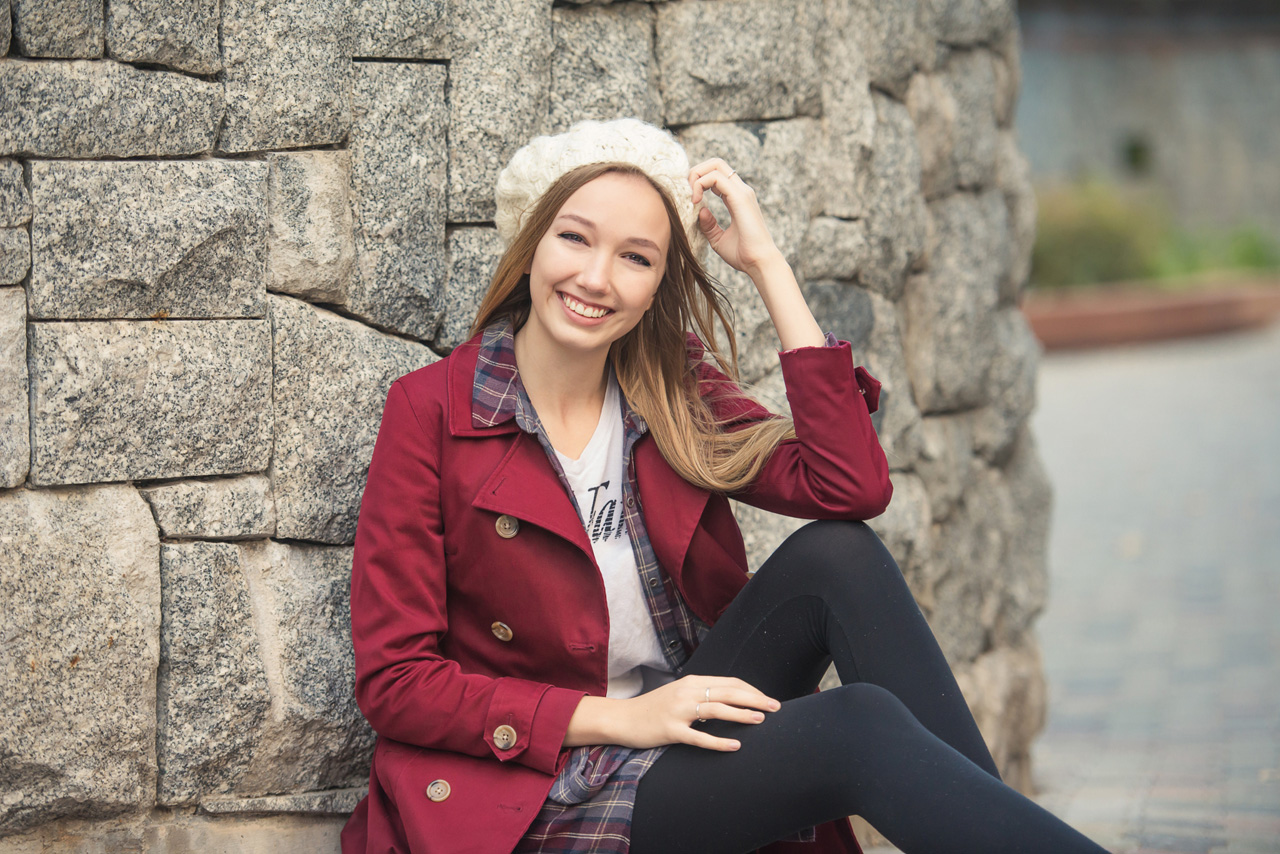 Girl in red pea coat sitting against a rock wall