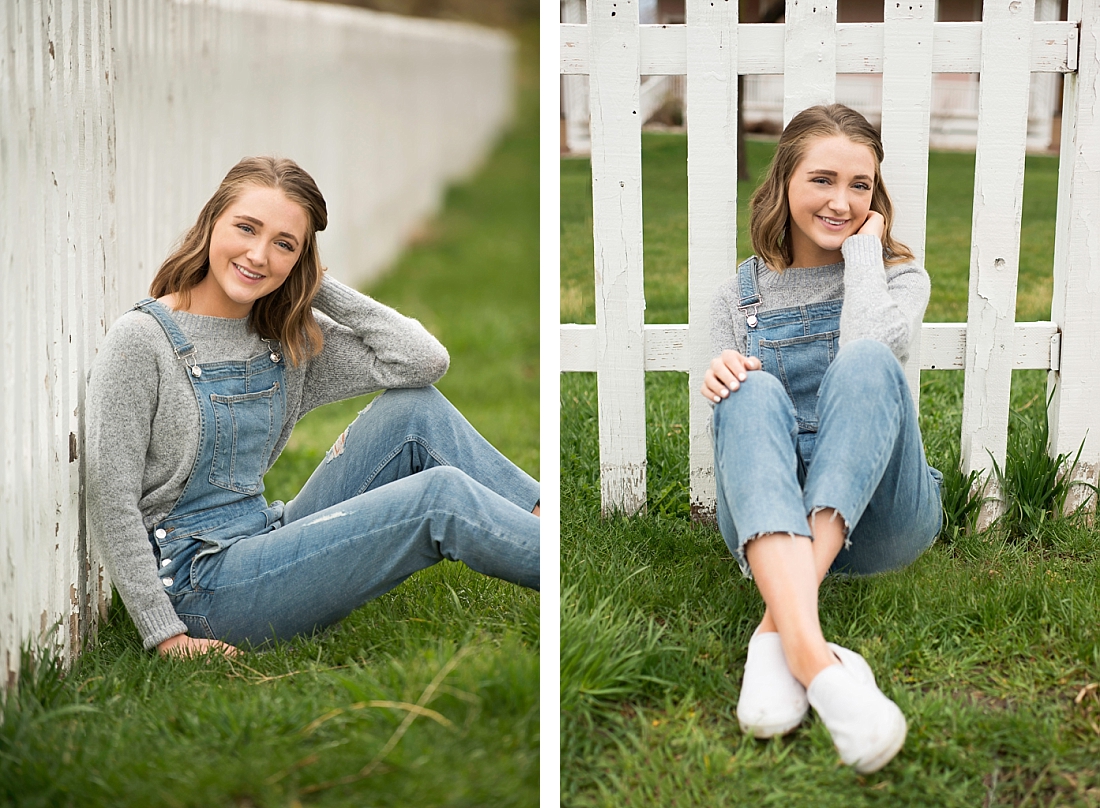Skyline High School senior portraits girl in gray sweater and overalls sitting by a white picket fence