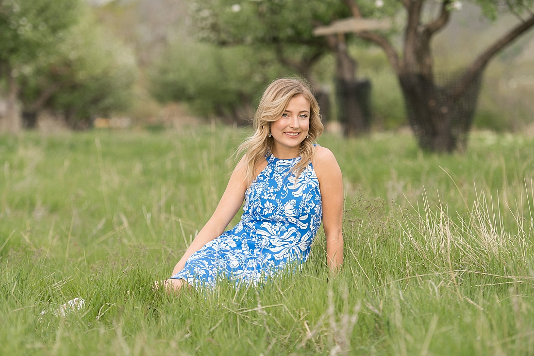girl in blue floral dress sitting in tall grass in apple orchard
