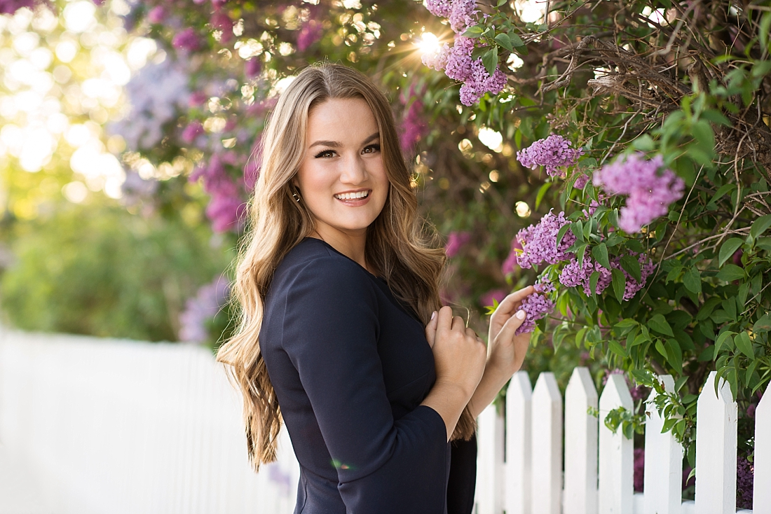 Amanda Nelson Photography Girl in navy blue dress standing by purple lilacs by a white picket fence