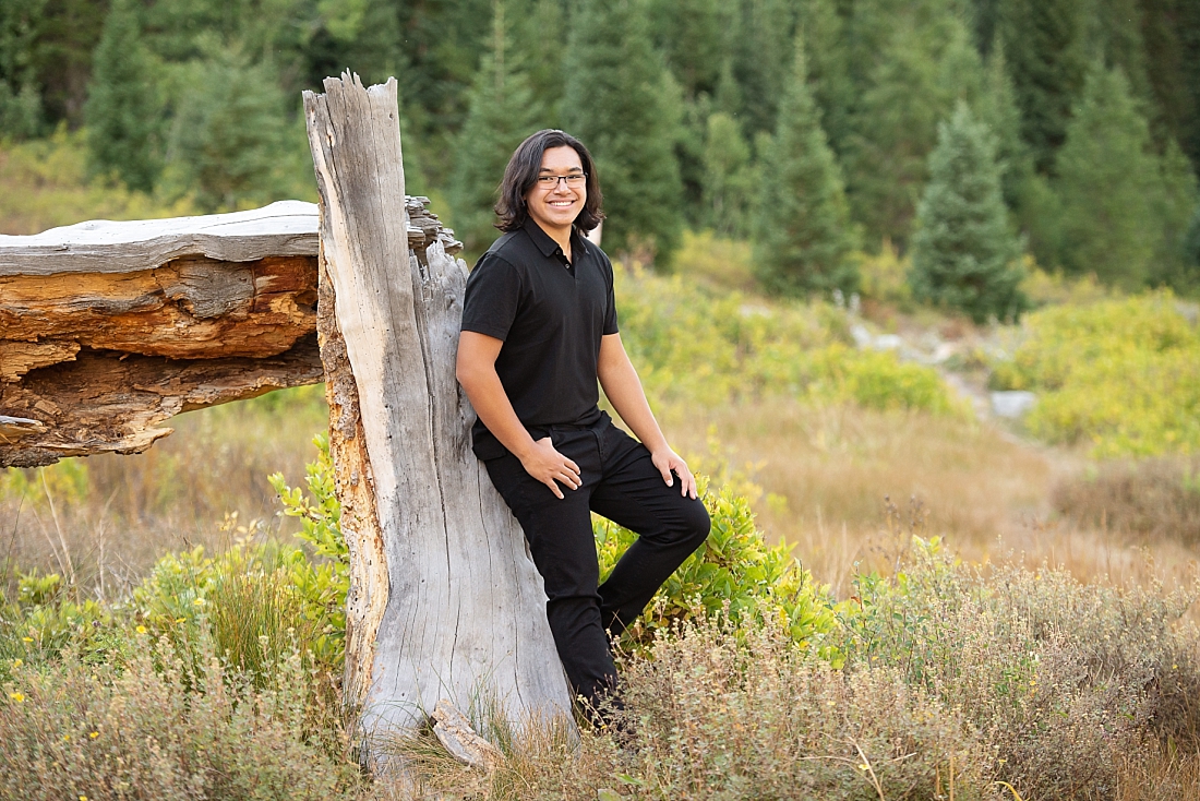 Waterford Senior Portraits in Utah mountains against a fallen tree. Black shirt and pants.