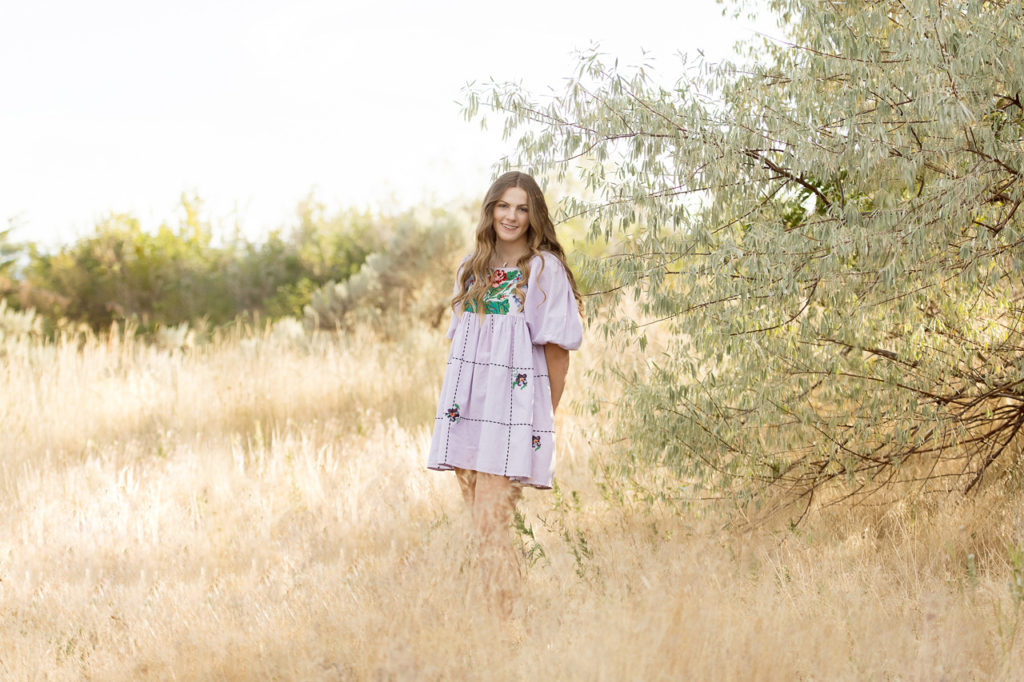 girl in lavender embroidered dress standing in wheat field by Russian olive tree