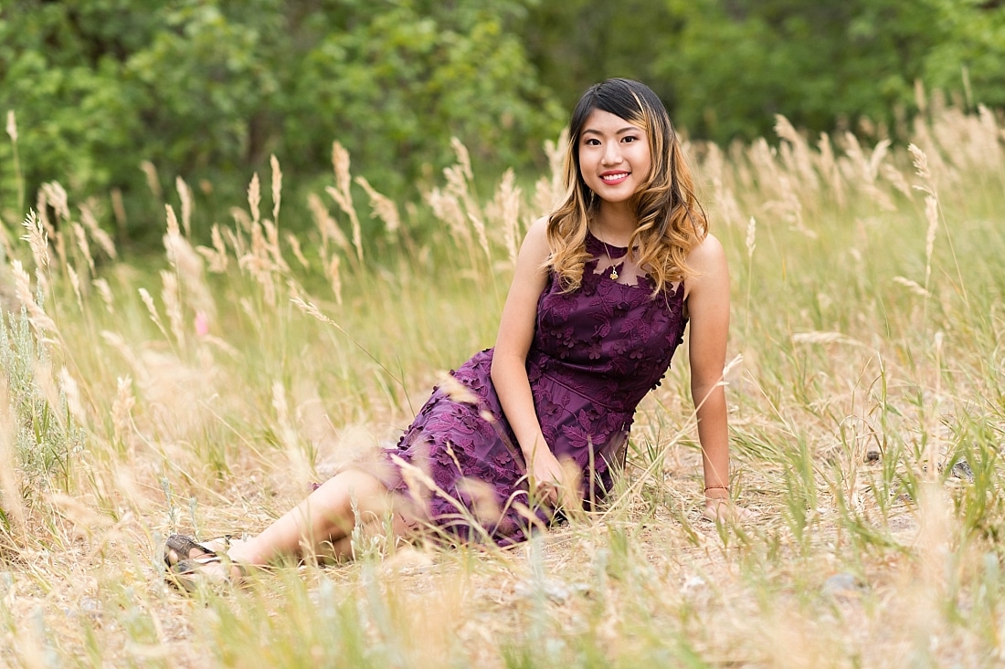 Girl sitting in the tall grass wearing a plum dress with lace overlay.