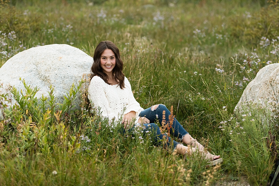 Waterford senior girl in cream sweater sitting against a rock in wild grass and flowers.