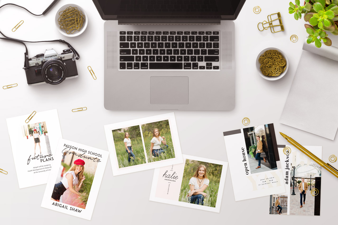 white table with laptop, coffee, and a camera holds three sets of high school graduation announcements.