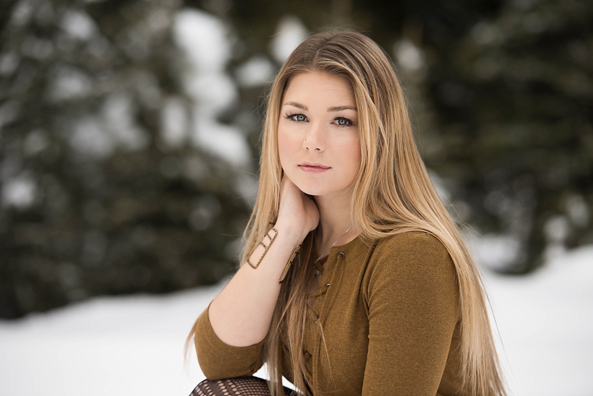 Girl in brown dress sitting in the snow with pine trees in the background
