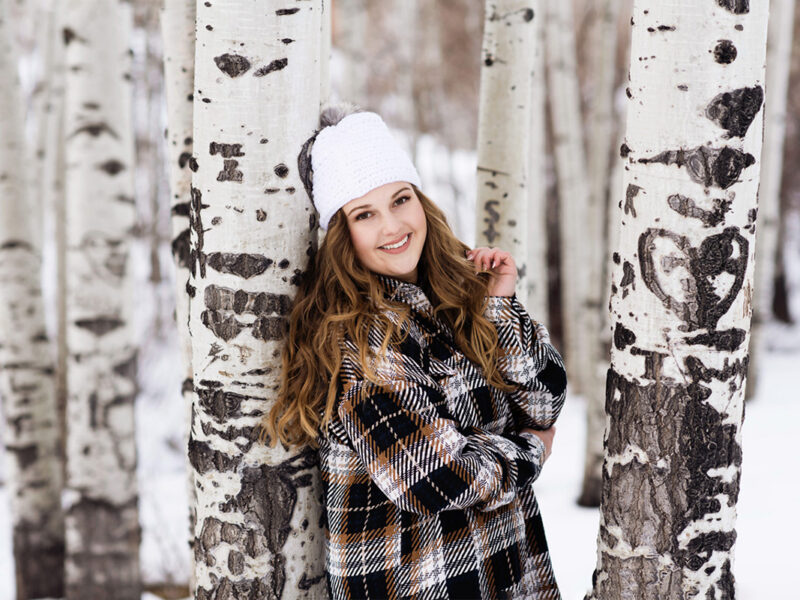 Girl in plaid button-up and beanie leaning against an aspen tree in the snow