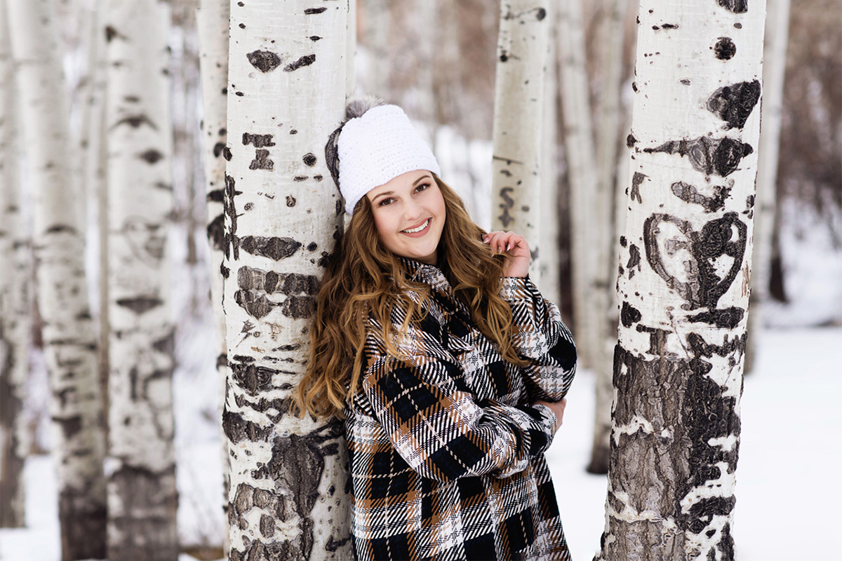 Girl in plaid button-up and beanie leaning against an aspen tree in the snow